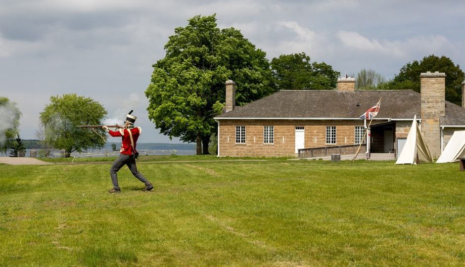 a costumed interpreter demonstrates black powder musket firing in front f the original Officers Quarters